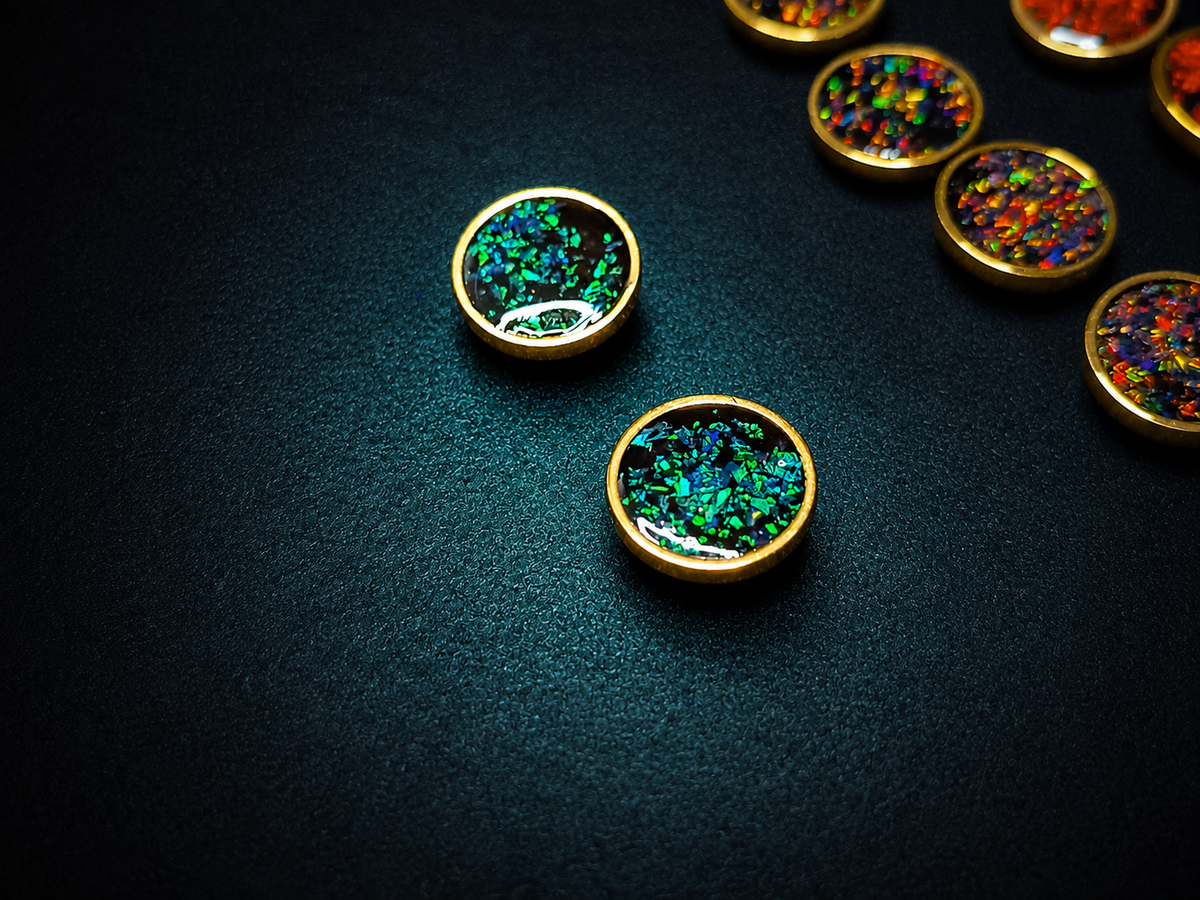 Gold-rimmed oval cabochons filled with iridescent multicolor glitter, displayed on a dark textured background with several more cabochons partially visible.