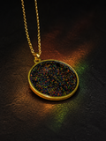 A gold pendant necklace with a round bezel containing a dense, iridescent multicolour glitter, resting on a dark textured surface with dramatic coloured lighting.