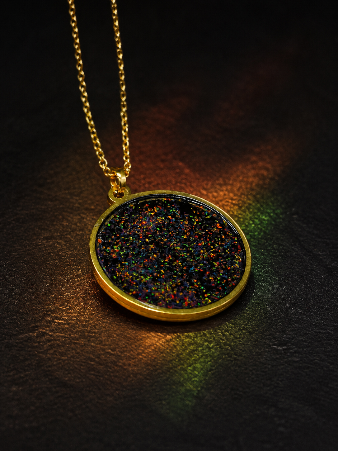 A gold pendant necklace with a round bezel containing a dense, iridescent multicolour glitter, resting on a dark textured surface with dramatic coloured lighting.