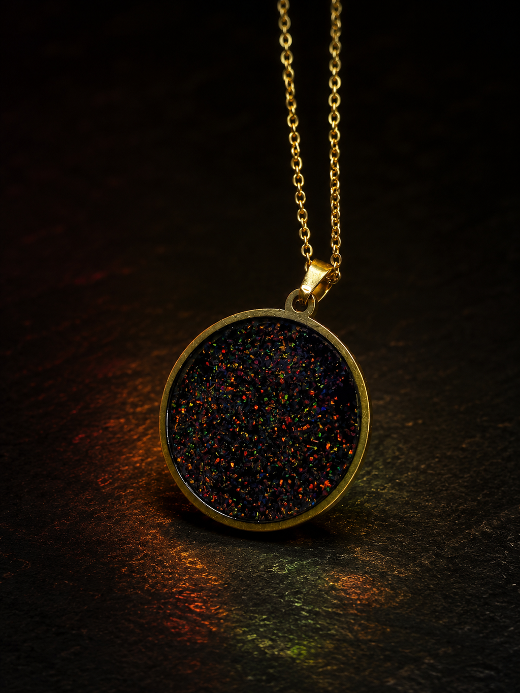 A gold pendant necklace featuring a round bezel set with a dark, glittery opal-like stone that shifts between black, red, and blue, hanging against a dark textured background.