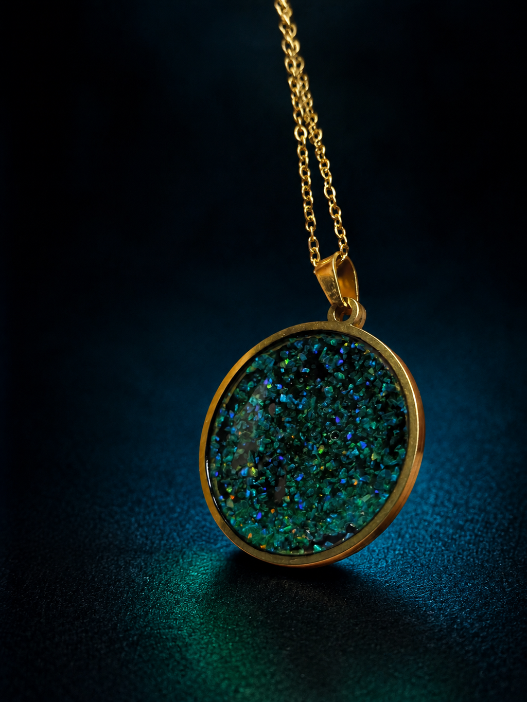 A gold pendant necklace featuring a round, glittery green and blue druzy stone, suspended from a gold chain against a dark, moody background.