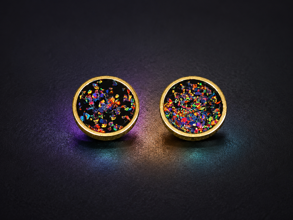 A pair of gold-tone stud earrings with round black centers filled with iridescent, rainbow-colored glitter, photographed on a dark textured background.