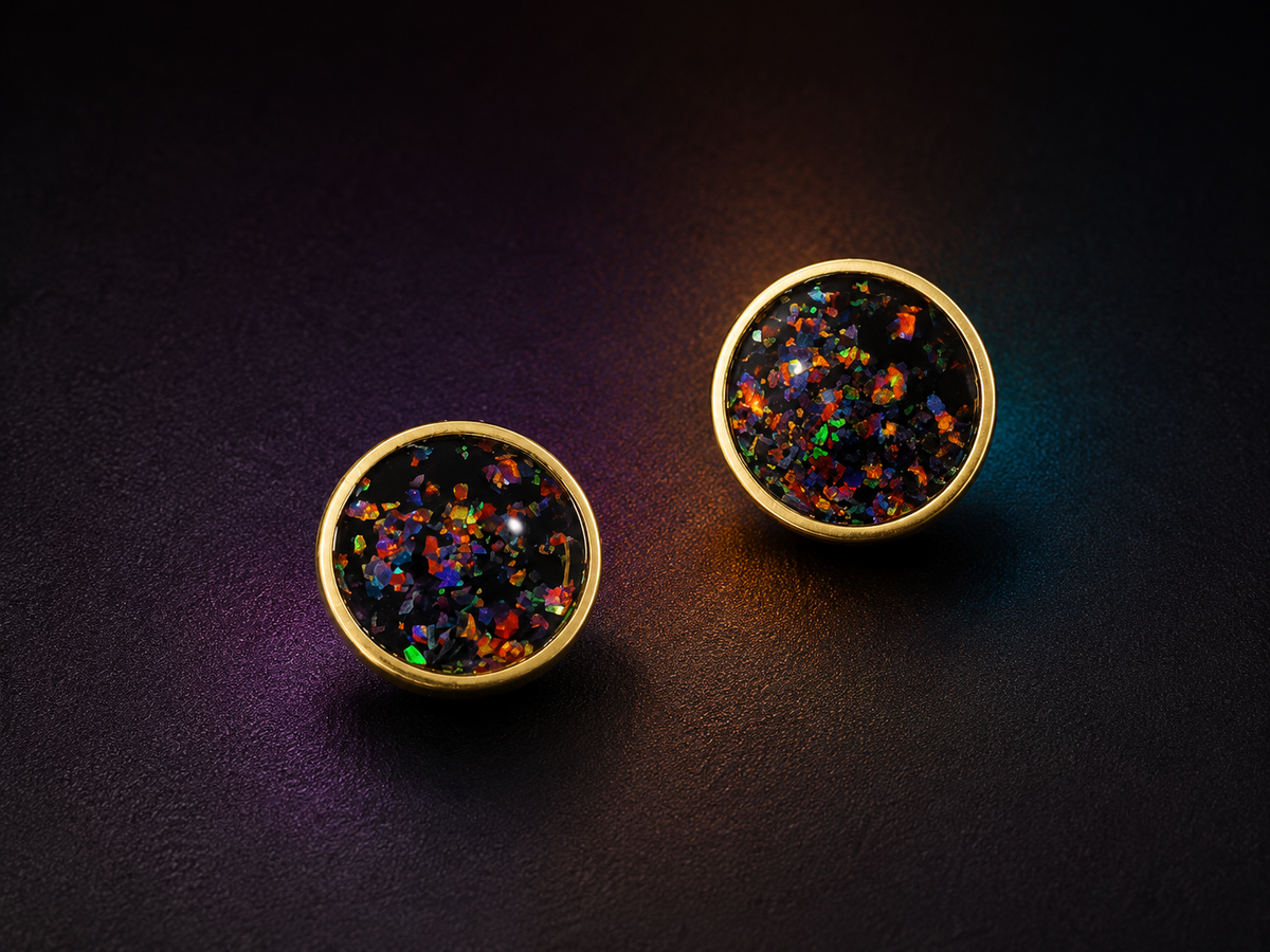 A pair of round gold-tone stud earrings with black resin tops filled with multi-coloured iridescent glitter, displayed on a dark textured background.